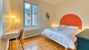 Un dormitorio con piso de madera, una cama blanca con cabecera naranja, un escritorio con una silla y una ventana que ofrece vistas a edificios.