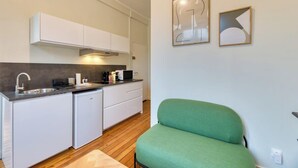 Una cocina compacta con armarios blancos, un microondas integrado y un lavamanos. Hay una silla verde y dos piezas de arte abstracto en la pared.