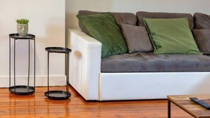 Un salon moderne avec un canapé blanc, des coussins verts et gris, deux petites tables d’appoint noires et une petite plante en pot.