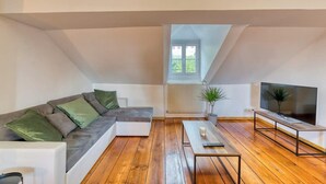 Un salon moderne avec un plancher en bois, un canapé d’angle gris agrémenté de coussins verts, un téléviseur à écran plat posé sur un meuble et un radiateur en arrière-plan.