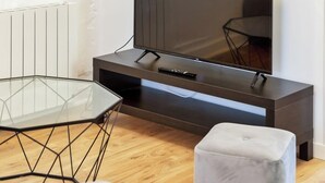A modern living room with a flat-screen TV on a dark wooden TV stand, a glass table with a geometric frame, and a light-colored ottoman. - Damien, votre *F3 *2 chambres *terrasse à *Rouen (Rouen)
