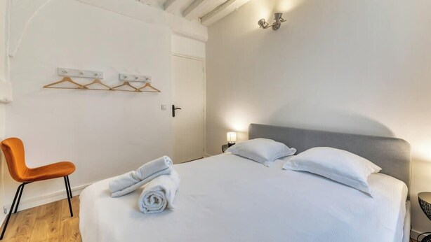 A neatly made bed with white linens and two pillows, a wooden chair, a wall-mounted light fixture, and a door with a handle. - Damien, votre *F3 *2 chambres *terrasse à *Rouen (Rouen)