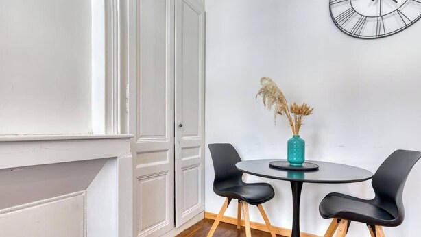 A room with a round table, two black chairs, a vase with dried plants, and a wall-mounted clock.