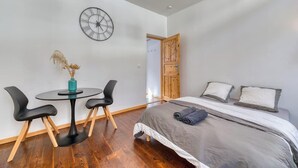 A bedroom with a bed, a round table with chairs, a clock on the wall, and a wooden door.