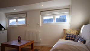A bedroom with a bed, two windows, a radiator, a wooden table, and a small table lamp. - Le Petit Nid * en plein centre-ville de Rouen ! (Rouen)