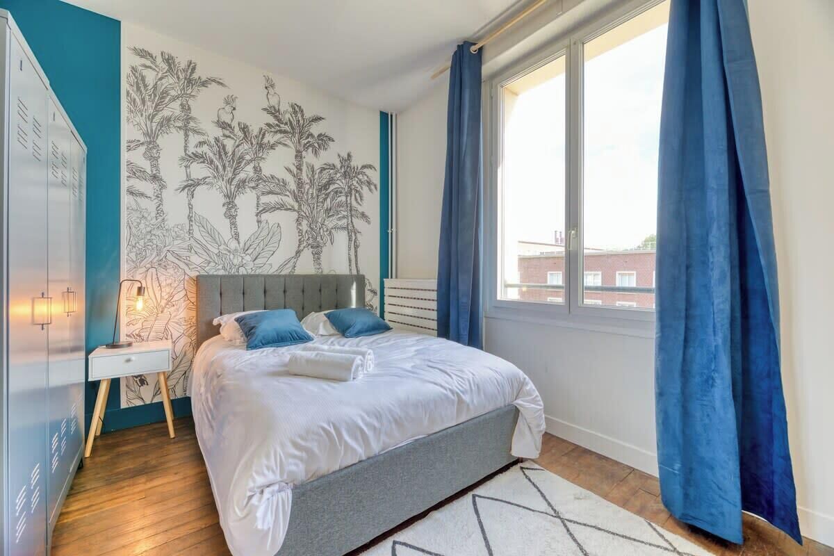 A bedroom with a bed, a nightstand, a wall mural, and a window with curtains.