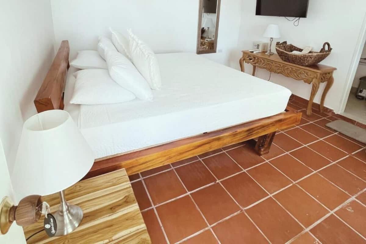 A bedroom with a wooden bed frame, white bedding, a bedside table with a lamp, a TV mounted on the wall, and a small decorative basket.