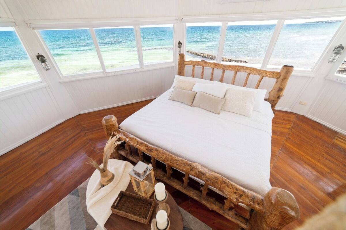 A wooden bed with white bedding in a room with large windows overlooking the sea.