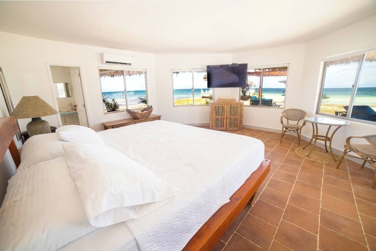 A bedroom with a large bed, a TV, a desk with a chair, and a view of the beach.