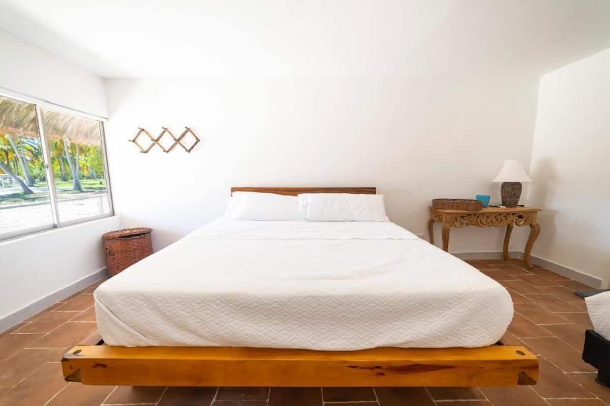 A bedroom with a bed, a window, a small table with a lamp, and a decorative wall piece.