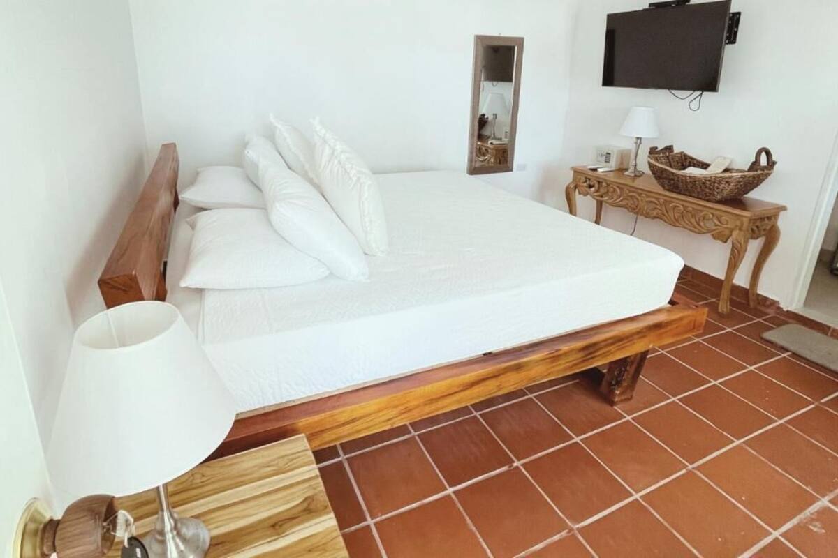 A bedroom with a wooden bed frame, white bedding, a bedside lamp, a television on the wall, and a small wooden table with a basket.