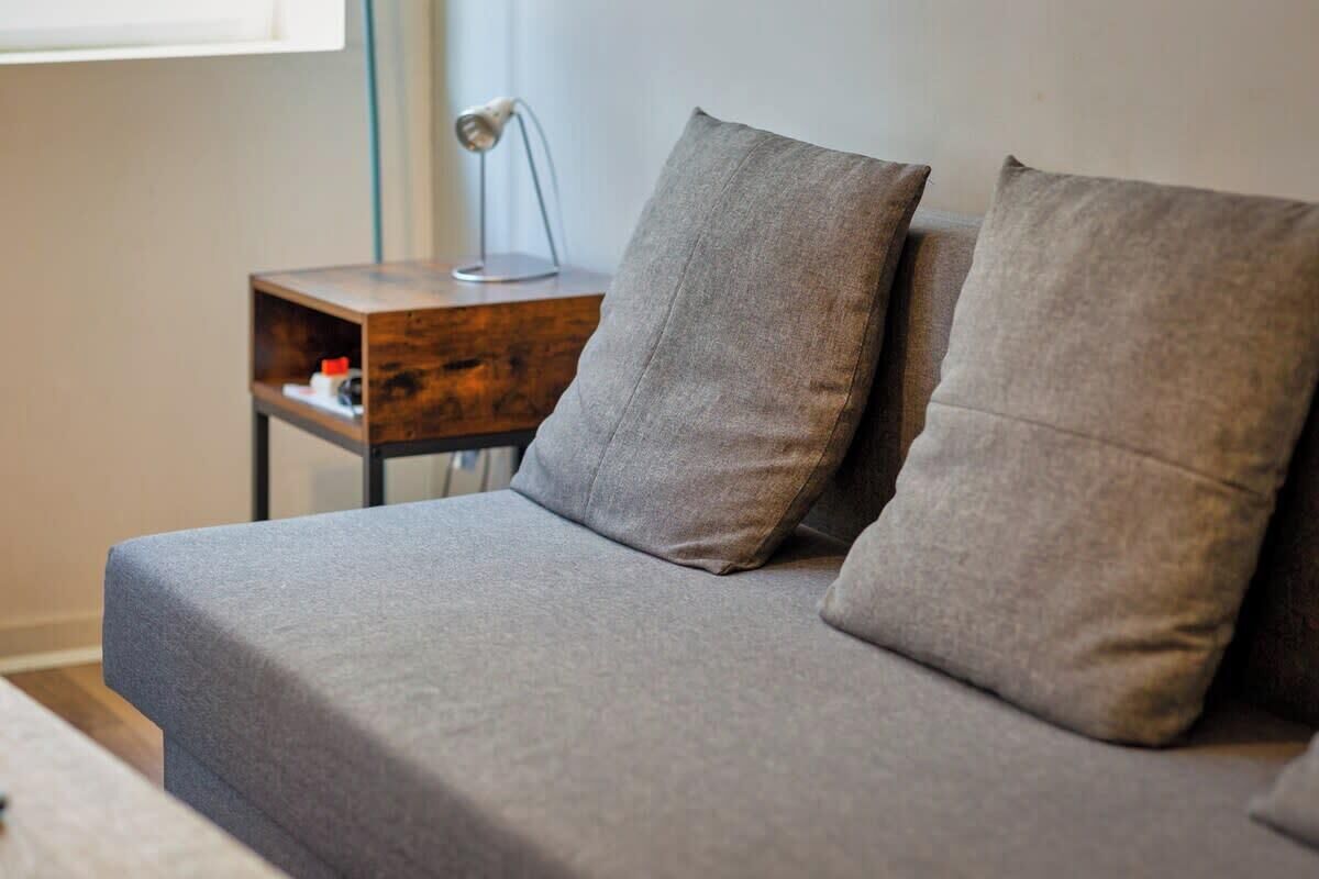 Ein Bett mit grauer Bettwäsche und zwei Kissen, ein Nachttisch aus Holz mit einer Lampe und eine kleine Ablage mit Gegenständen.