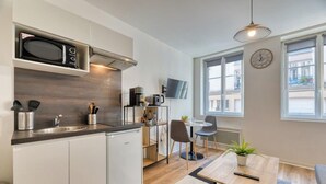 Una cocina compacta equipada con microondas, cafetera y un refrigerador pequeño. Hay un comedor con mesa y sillas, y una ventana con vista a la calle.
