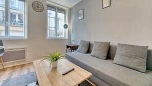Un salón moderno con un sofá gris, una mesa de centro de madera con una planta, un reloj en la pared y ventanas amplias.