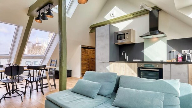 Een modern loft appartement met een groot dakvenster, een kitchenette, een eethoek en een lichtblauwe bank.