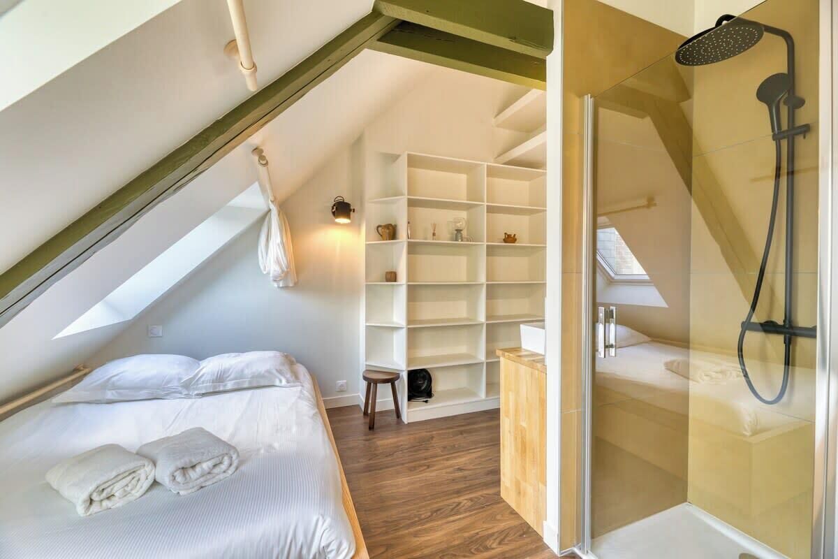 Une chambre avec un lit, un petit tabouret, une étagère et un espace douche.