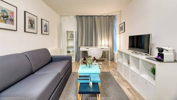 En moderne stue med en grå sofa, et blåt sofabord, et fjernsyn og et hvidt skrivebord med en computer.