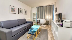 En moderne stue med en grå sofa, et lille blåt sofabord og et skrivebord med en computer. Væggene er dekoreret med billeder, og der er et tv på et stativ.