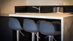 Une cuisine moderne avec un plan de travail blanc, un seul robinet et trois tabourets de bar. On y trouve trois récipients gris étiquetés « SUCRE », « THÉ » et « CAFÉ ».