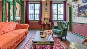 En hyggelig stue med sofa, lænestol og sofabord. To vinduer med gardiner, en væglampe og et TV ophængt over sofaen.