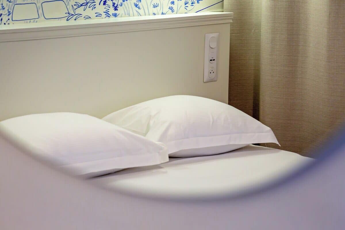 A bed with white bedding in a room with a white wall and a power outlet.