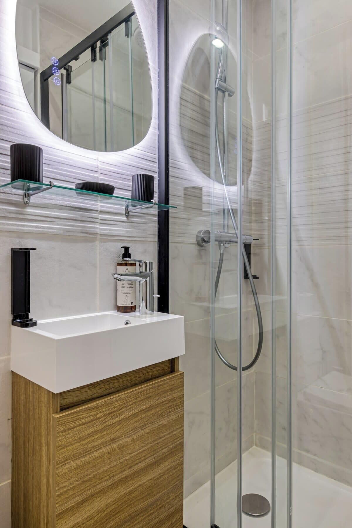 Un baño moderno con ducha envuelta en vidrio, tocador blanco con gabinete de madera y un espejo grande de marco circular.