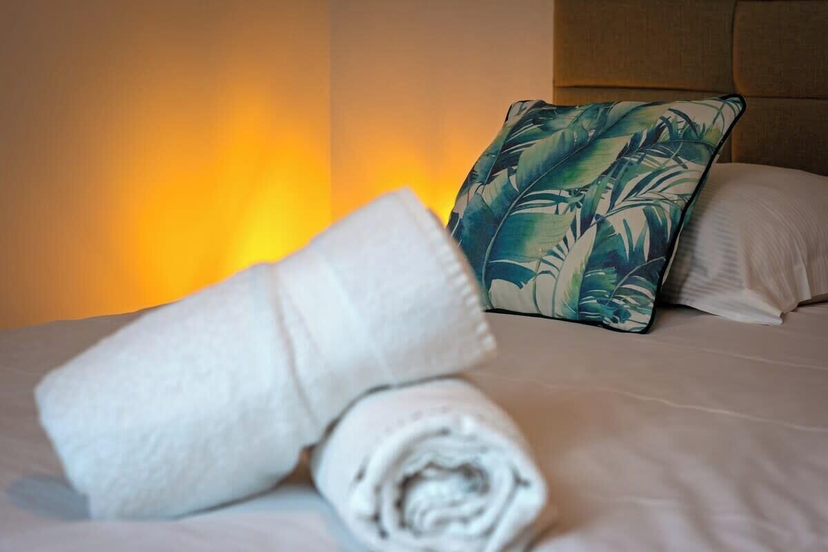 Un lit bien fait, avec une serviette blanche roulée et un oreiller orné d’un motif de feuilles tropicales.