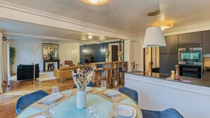 A modern dining area with a round table set for four, a kitchen in the background, and a living area with a sofa and a lamp.