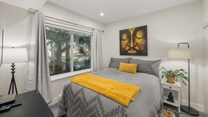 A bedroom with a bed, a window with curtains, a painting on the wall, a bedside lamp, and a small table with a plant. - Stunning waterfront apartment - wake up and fall asleep listening to the ocean (Delta)
