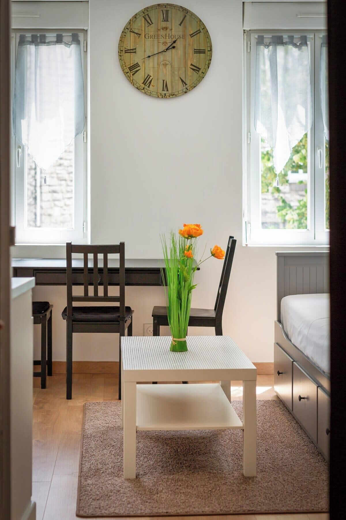 A small room with a wooden chair, a white table, a clock on the wall, and a window with curtains.