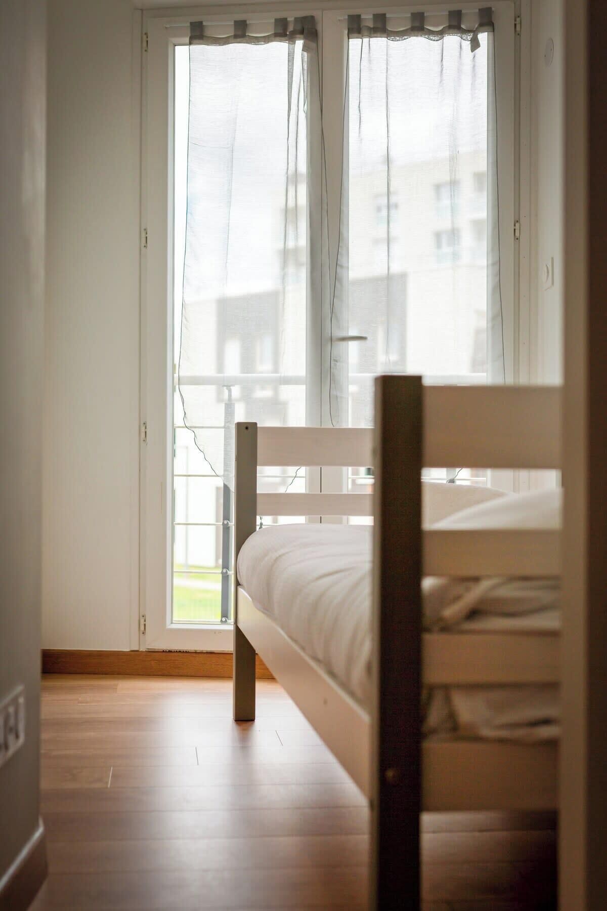 A bedroom with a wooden bed, a window with sheer curtains, and a view of buildings outside.