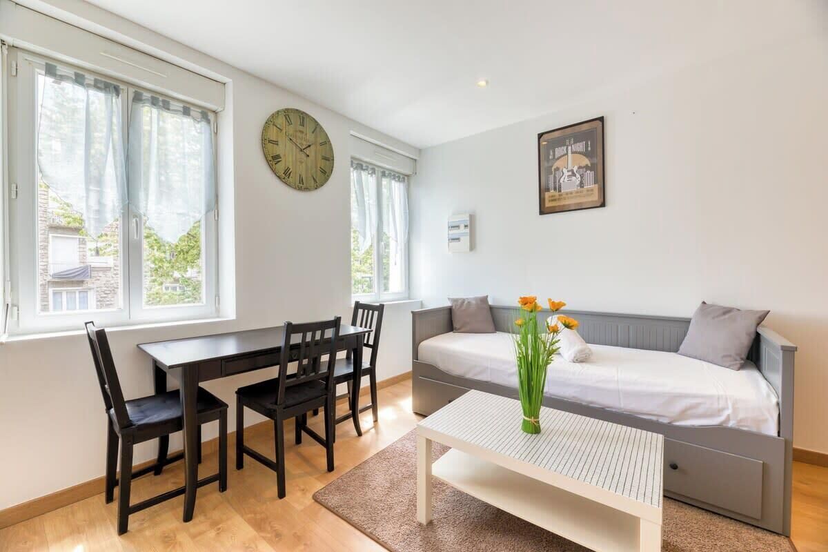 A room with a bed, a desk, and chairs. There is a clock on the wall and a picture frame above the bed.