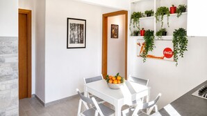 Zona pranzo con tavolo bianco, sedie, una ciotola di frutta e uno scaffale a parete con piante e un cartello Coca-Cola.