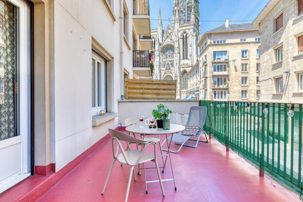 A balcony with a red floor, two chairs, a table with a plant, and a view of a cathedral.