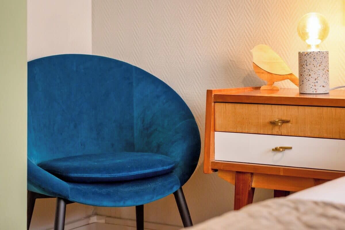 Une chaise bleue est placée à côté d’une table de chevet en bois, sur laquelle se trouvent une lampe et un objet décoratif.