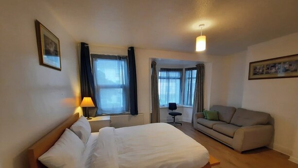 Habitación de hotel con una cama, un escritorio, una silla, un sofá y dos ventanas con cortinas.