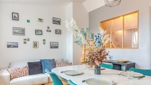 En moderne stue med et spisebord, turkise stole, en hvid sofa med mønstrede puder og en væg pyntet med billeder i rammer.
