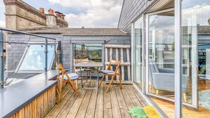 En moderne udendørs terrasse med trægulv, glasvægge og et siddeområde med stole og et bord.