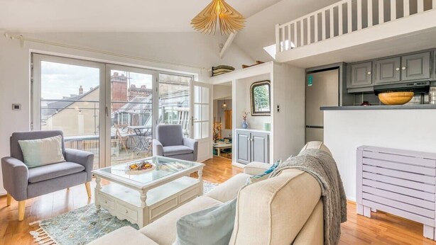 En rummelig stue med et sofabord i glas, to lænestole og en sofa. Et køkkenområde med et moderne design ses i baggrunden.