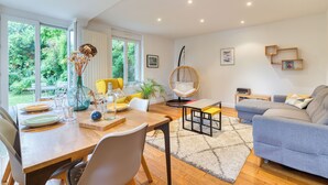 A modern living room with a dining area, a sofa, a hanging chair, and a view of greenery outside.