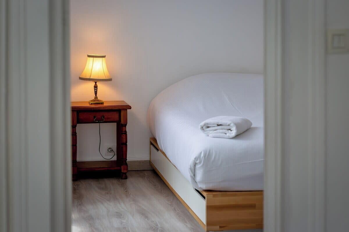 Ein ordentlich bezogenes Bett mit weißer Decke und gefaltetem Handtuch, eine Nachttischlampe auf einem Holznachttisch und Holzboden.