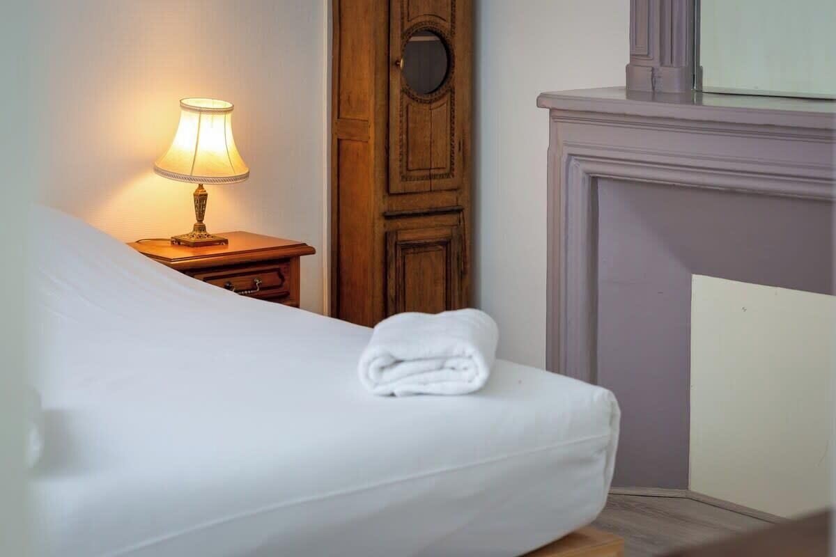 Una cama bien tendida con una toalla blanca doblada, una mesita de noche con una lámpara y una puerta de madera con un espejo redondo.