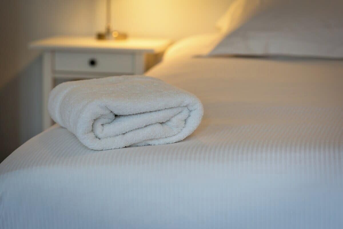 A neatly made white towel on a bed with a white headboard and a bedside table with a lamp.