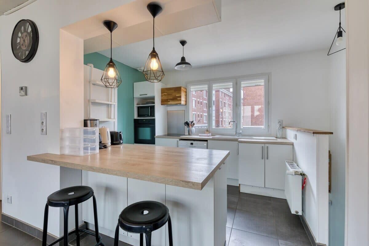 A modern kitchen with a central island, two bar stools, pendant lighting, and a view of the city through large windows.
