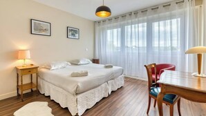 A hotel room with a bed, a wooden table, a chair, a lamp, and a window with curtains.