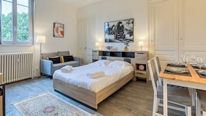 Ein modernes Schlafzimmer mit einem großen Bett, Nachttischen, einem Schreibtisch mit Stühlen, einem Heizkörper und einem Fenster mit Blick auf Bäume.