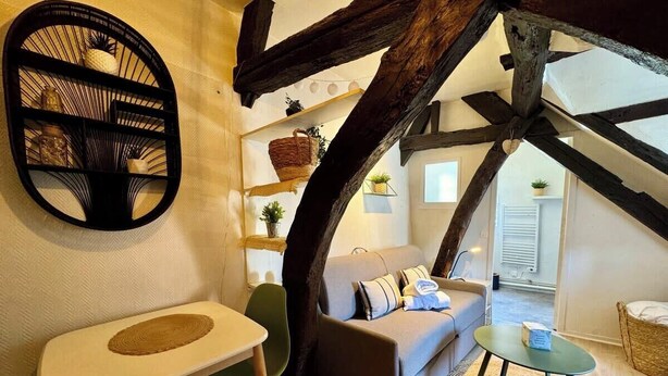 A cozy loft-style living space with exposed wooden beams, a sofa, a small table, and a wall-mounted shelf with decorative items. - Vera : Studio Confortable sous les combles ! (Rouen)
