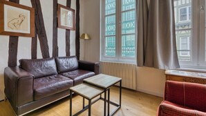 A room with a brown leather sofa, a small wooden table, and a red checkered chair. There are framed pictures on the wall and a window with curtains.
