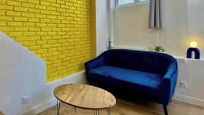 Een kamer met een geel bakstenen muur, een blauwe bank, een ronde houten tafel en een raam met gordijnen.