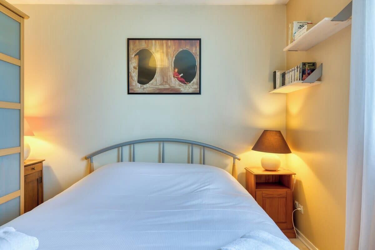 A bedroom with a bed, a nightstand with a lamp, a wall-mounted shelf with books, and a painting above the bed.